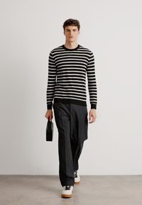 Sweater a rayas en blanco y negro con cuello redondo, combinado con pantalones negros ajustados y zapatillas blancas. Sostiene un pequeño bolso negro.