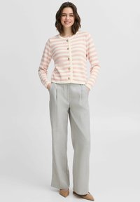 Svītrains kardigans rozā un krēma krāsā ar sešām zelta pogām, pieguļošs dizains, garas piedurknes, kombinēts ar gaišiem pelēkiem biksēm un bežām kurpēm ar papēžiem.