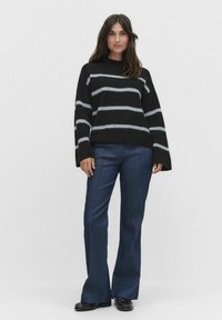 Svart och grå randig tröja med långa ärmar, i kombination med mörkblå utsvängda byxor och svarta skor, mot en ljus bakgrund.