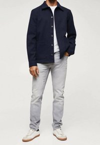 Chaqueta navy con botones y cuello, dos bolsillos frontales, combinada con jeans de un azul claro y zapatillas blancas con acentos marrones.