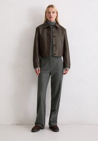 Braune Lederjacke mit kurzem Schnitt, Kragen, Frontknöpfen, einem grauen Rollkragenpullover darunter und grauen gemusterten Hosen. Dunkle Schuhe.