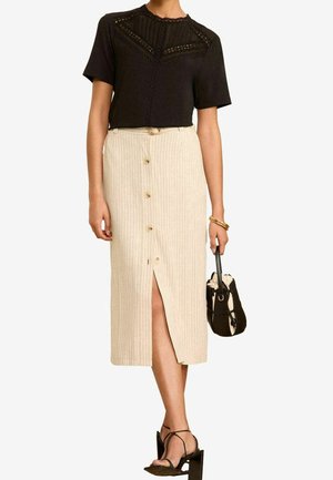 Vrouw draagt een zwart topje met kanten details, een beige midi-rok met knopen en een split, zwarte sandalen met bandjes, een gouden armband, en houdt een zwart-beige handtas vast.