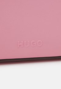 Rosa faux skinnmateriale med preget "HUGO"-logo. Glatt tekstur med minimal detaljering og svart kant nederst.