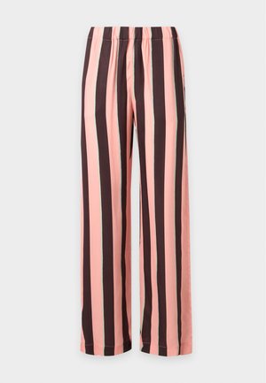 Femme portant une chemise rouge à manches courtes, un pantalon large à rayures verticales roses et marron, et des chaussures plates noires, debout devant un fond uni.