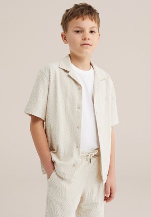 Fiú, aki texturált krémszínű rövid ujjú gombos ingben és hozzáillő rövidnadrágban van, zsebre tett kézzel, egyszínű háttér előtt.