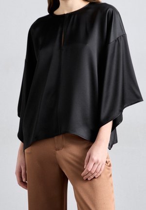 Persoon die een losse zwarte blouse met brede mouwen en een bruine broek draagt, staand tegen een effen lichtgrijze achtergrond.