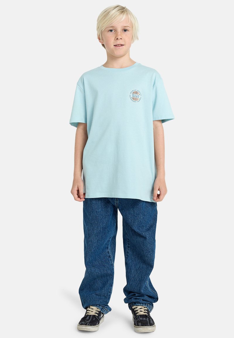 T-shirt di cotone azzurro chiaro con scollatura rotonda e maniche corte, abbinata a jeans di denim scuri e larghi con texture e sneakers nere.