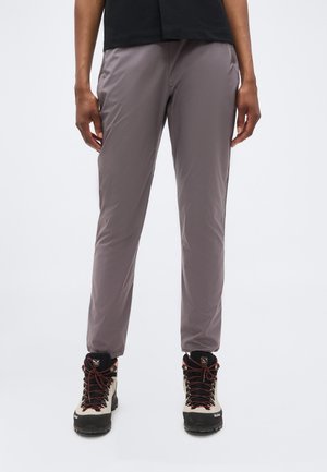 Osoba koja nosi sive sužene hlače i bež planinarske čizme s crnim potplatima i crvenim vezicama, stoji na bijeloj pozadini.