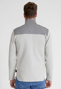 Grau Fleece-Pullover mit dunkelgrauem Schulterpanel, Stehkragen und langen Ärmeln, kombiniert mit blauen Jeans. Glatte Textur und lässiges Design.