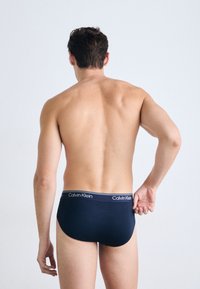 Mann mit kurzen dunklen Haaren, von hinten gesehen, trägt navyblaue Calvin Klein Slip, zieht mit der rechten Hand den Bund gegen einen schlichten Hintergrund.