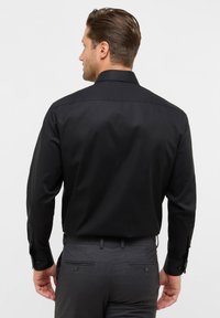 Zwarte langarmshirt met een gladde textuur, klassieke kraag en met knopen sluitende manchetten, gedragen met een grijze broek met achterzakken.