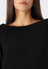 Fekete, textúrált felső széles nyakkivágással. Az anyag steppelt mintázatot mutat, amely mélységet és vizuális érdekességet ad a dizájnnak.