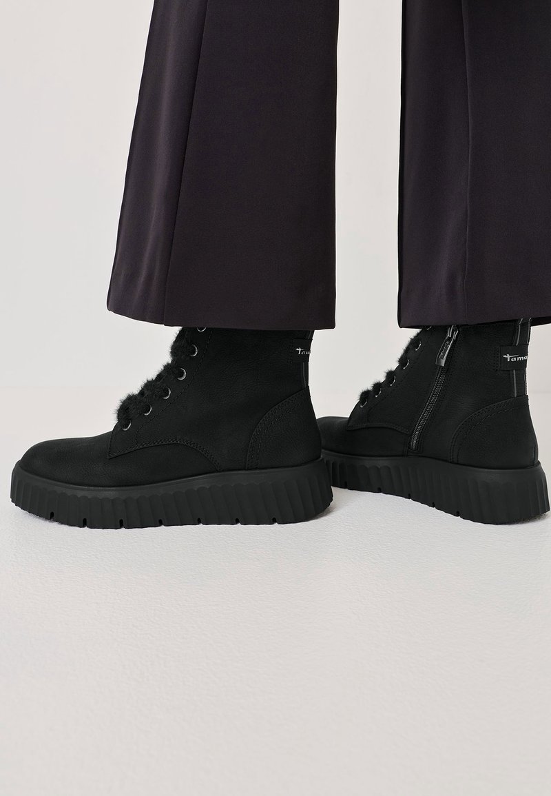 Chevilles portant des bottes noires en suède à lacets, avec des semelles épaisses rainurées et des fermetures éclair latérales, associées à un pantalon large et foncé.