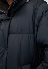 Sort pufferjakke med høj krave, der har et kviltet design, metal lynlås og en sidelomme. Glat, blank overflade.