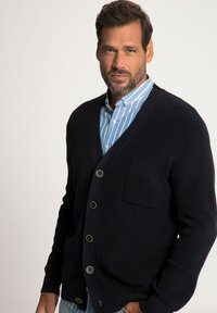 Cardigan a costine blu navy con cinque bottoni, caratterizzato da una tasca sul petto e indossato sopra una camicia a righe azzurra.