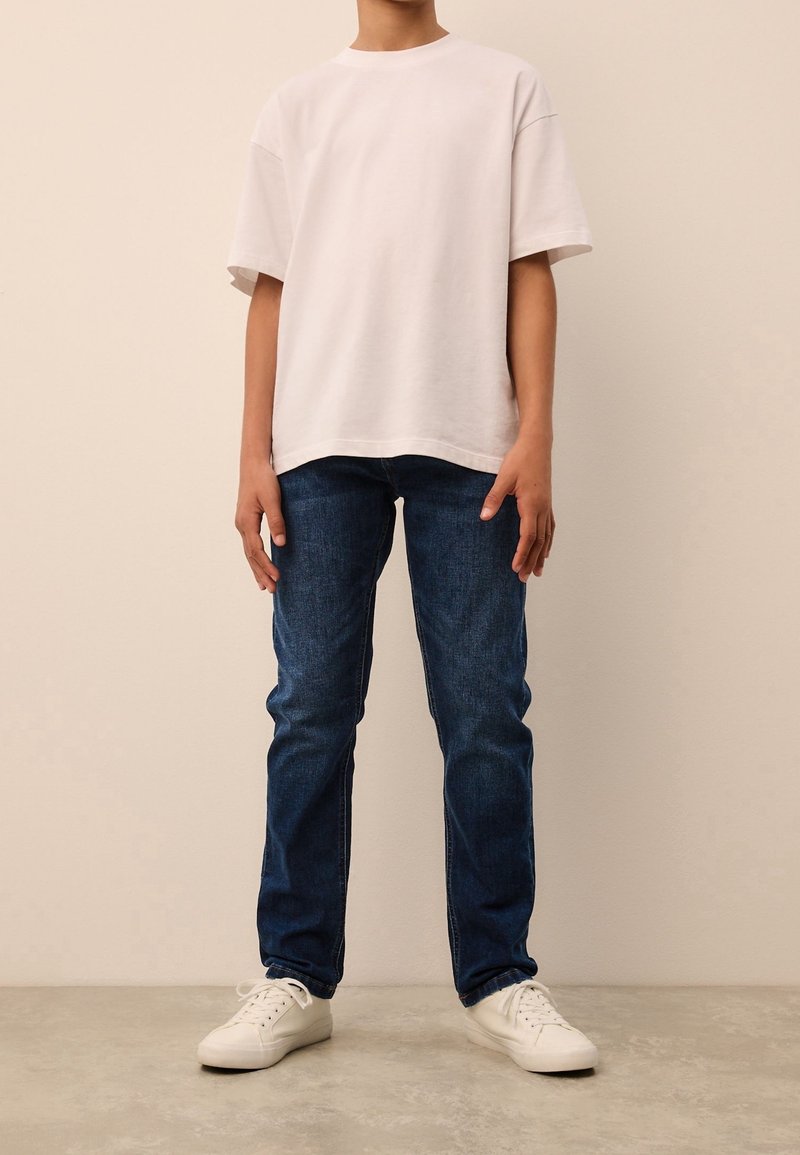 Personne debout portant un t-shirt blanc ample à manches courtes, un jean bleu foncé et des baskets blanches sur un fond clair uni.