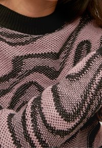 Pull en maille rose avec des motifs noirs ondulés, doté d'un col côtelé noir. La texture semble douce et respirante.