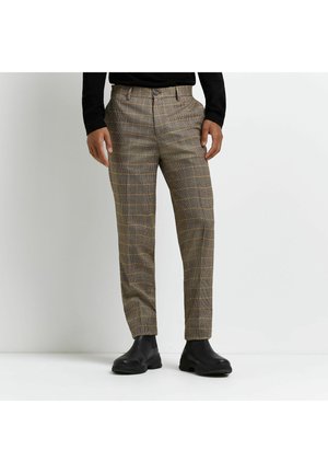 Man draagt bruine geruite broek, zwart shirt met lange mouwen en zwarte leren enkellaarsjes, staand tegen een witte muur.