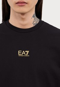 Fekete pamut póló kerek nyakkal, arany EA7 Emporio Armani logóval a bal mellrészen. Puha tapintású, testhez simuló fazon.
