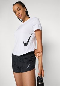 Biele tričko Nike s veľkým čiernym logom swoosh, v kombinácii s čiernymi športovými šortkami obsahujúcimi malé biele logo swoosh, držané v jednej ruke s fľašou vody.