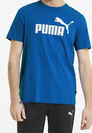 Personne portant un t-shirt Puma bleu avec un logo blanc et un pantalon noir arborant également un petit logo Puma blanc.