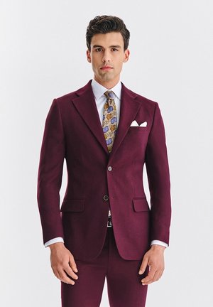 Bordowy garnitur o dopasowanym kroju, z klapami z wcięciem i dwoma przednimi kieszeniami. Do tego krawat w wzory z niebieskimi i złotymi akcentami.