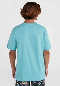 Una camiseta de manga corta de color azul claro con cuello redondo, hecha de suave tela de algodón, mostrada en una persona que lleva puesto un bañador de estampado.