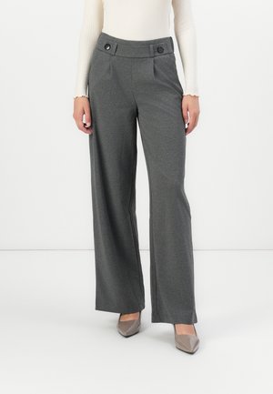Vrouw draagt grijze hoge taille broek met wijde pijpen en een met knopen afgewerkte tailleband en beige puntige hakken, staand tegen een witte achtergrond.