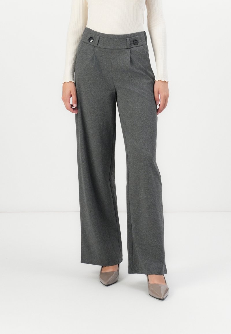 Femme portant un pantalon gris taille haute à jambes larges avec une ceinture boutonnée et des talons beiges à bouts pointus, debout devant un fond blanc.