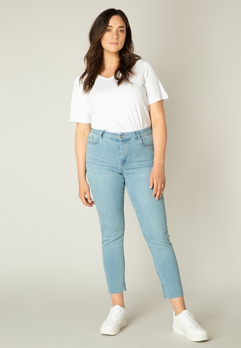 Lichtblauwe skinny jeans met een ruwe zoom, gecombineerd met een plain wit T-shirt en witte platform sneakers, staande tegen een neutrale achtergrond.