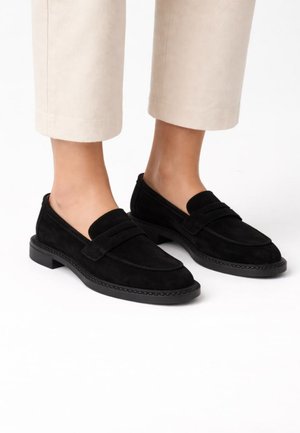 Mocassins en daim noir portés avec un pantalon court beige clair sur fond blanc, affichant un style épuré et minimaliste.