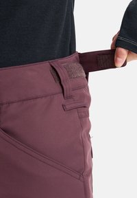 Persoon die de verstelbare tailleband met klittenbandsluiting van paarse broek aanpast, terwijl hij een donker T-shirt met lange mouwen draagt.