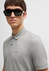 Gaiši pelēks polo krekls ar īsām piedurknēm, pogu slēgšanu un logotipa detaļu. Melni saulesbrilles ar biezām rāmī un sudraba akcentiem.