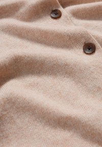 Weiches, strukturiertes Gewebe in einem hellen Pfirsichton mit zwei dunkelbraunen Knöpfen, das subtile Strickmuster und sanfte Drapierungen zeigt.