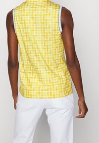 Gul rutigt ärmlöst linne med vita accenter, med ett schackmönster och en slät textur, bärs tillsammans med vita byxor.