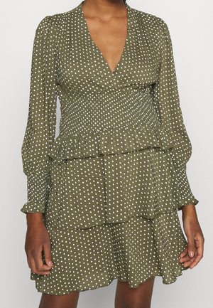 Olivgrünes Kleid mit weißen Punkten, langen Puffärmeln, geraffter Taille, V-Ausschnitt und mehrstufigem Rüschenrock an einer Person.