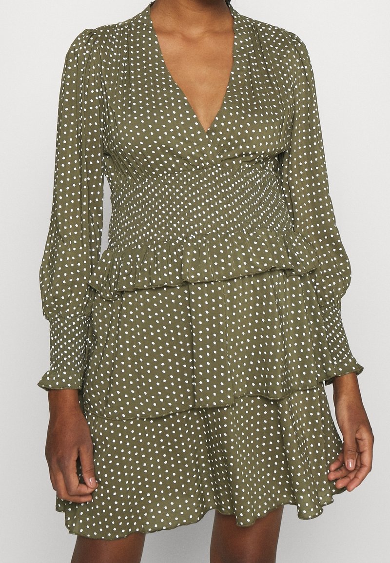 Robe vert olive à pois blancs, manches longues bouffantes, taille smockée, décolleté en V et jupe à volants à étages portée par une personne.