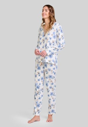 Mujer de pie descalza, vistiendo un conjunto de ropa de casa blanco con estampado floral azul, sonriendo con las manos entrelazadas al frente.
