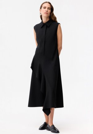 Vrouw met golvend haar die een mouwloze zwarte midi-jurk draagt met asymmetrische ruches, kraag en zwarte loafers, staande tegen een witte achtergrond.