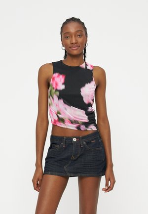 Jeune femme portant un crop top noir sans manches à motif floral rose et une mini-jupe en jean foncé, debout devant un fond blanc uni.