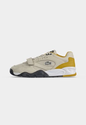 Beżowy niski sneaker z czarno-białą podeszwą, żółtym akcentem na pięcie, sznurowany z paskiem na rzep i logo krokodyla Lacoste z boku.