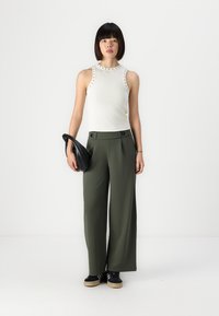 Witte mouwloze top met studaccenten, gecombineerd met hoge, wijde olijfgroene broeken. Houdt een zwarte clutch vast, staande op een effen achtergrond.