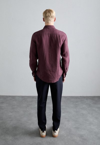 Camisa de lino burdeos de manga larga con dobladillo curvado y puños de botón, combinada con pantalones oscuros y zapatos claros con suela texturizada.
