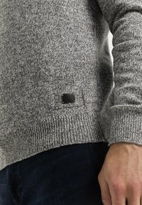 Grauer Strickpullover mit strukturiertem Muster, ausgestattet mit einem schwarzen Etikett am unteren Saum. Weicher Stoff mit gerippten Bündchen und Saum.