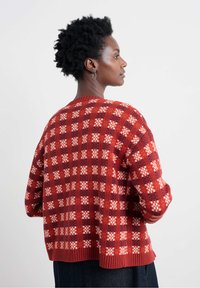 Žena s krátkymi kučeravými vlasmi, oblečená v pletenom svetri s červenými a oranžovými kockami, stojí v profile proti plain bielej pozadí.