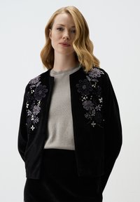 Cardigan in velluto nero con ricami floreali in viola e argento, caratterizzato da un scollo rotondo e frontale aperto, indossato su una maglia grigia.