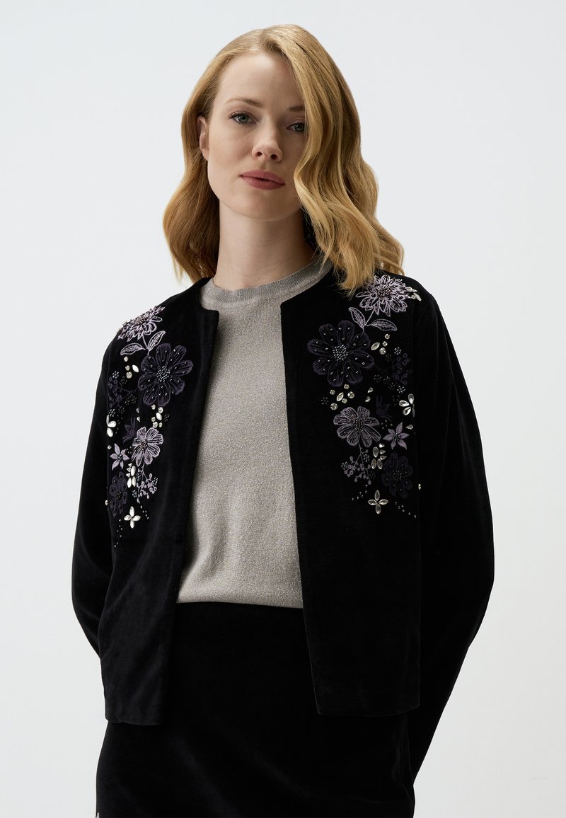 Cardigan in velluto nero con ricami floreali in viola e argento, caratterizzato da un scollo rotondo e frontale aperto, indossato su una maglia grigia.