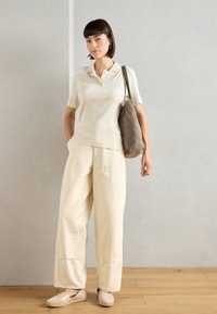 Mujer de pie con una polo de manga corta con relieve, pantalones amplios crema, zapatos beige de slip-on y un bolso de hombro taupe sobre un suelo de madera.