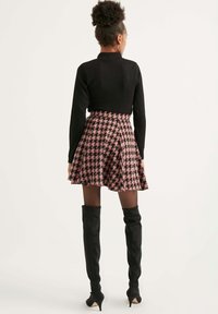 Schwarzes Langarmoberteil kombiniert mit einem rosafarbenen Hahnentrittmuster-gefalteten Rock. Das Modell trägt schwarze Stiefel mit Absatz, die über das Knie reichen.