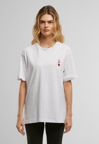 Bílý bavlněný tričko s krátkými rukávy a kulatým výstřihem, na levé hrudi má malou červenou bezpečnostní špendlík a design srdce.